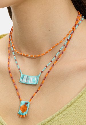 Trzy warstwowe naszyjniki z koralików w kolorze pomarańczowym, turkusowym i bordowym, z jednym wisiorkiem z napisem "JUICY" i drugim z okrągłym pomarańczowym wzorem.