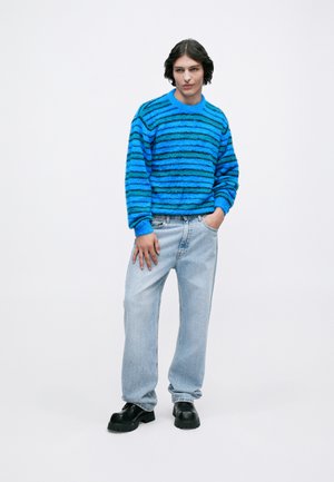 Pullover de malha com riscas azuis e tecido texturizado, combinado com jeans azul claro e sapatos pretos robustos; modelo está de pé com uma mão no bolso.