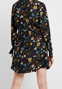 Mörkblå blommig klänning med cinchmidja, långa ärmar med knytningar och en livlig mix av vita, orange och turkosa blommor.