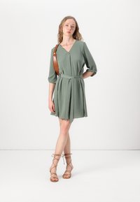 Vestido verde com mangas três quartos, cintura cingida e tecido texturizado. Combinado com sandálias de tiras cor de camelo contra um fundo neutro.