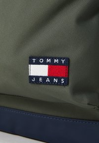 Green fabric bag featuring a navy base; includes a navy, white, and red embroidered patch displaying "TOMMY JEANS." Smooth, durable texture.