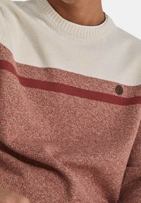 Roter und cremefarbener Strickpullover mit strukturiertem Muster, einer horizontalen kastanienbraunen Streife und einem kleinen Emblem auf der Brust.