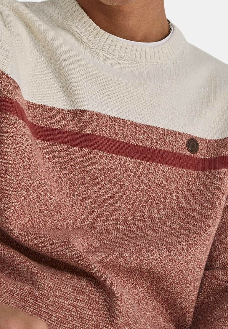 Roter und cremefarbener Strickpullover mit strukturiertem Muster, einer horizontalen kastanienbraunen Streife und einem kleinen Emblem auf der Brust.