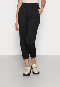 Kvinna i svarta högmidjade cropped-byxor med sidofickor och beige loafers med kraftiga svarta sulor, stående på ett grått golv.