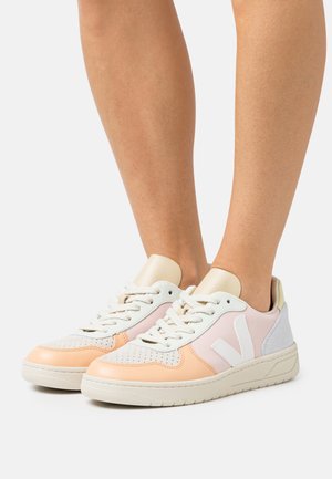 Beine, die flache Sneakers mit weißen, rosa und pfirsichfarbenen Paneelen, weißen Schnürsenkeln und beigen Sohlen vor einem schlichten Hintergrund tragen.