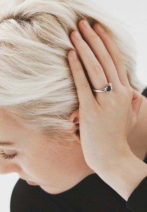 Anillo de plata con una piedra negra rectangular, llevado en una mano que toca el pelo corto y rubio claro. El fondo es suave y difuso.