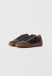 Adidași cu glezna joasă, din piele neagră și panouri din suede maro, cu tălpi din cauciuc, șireturi negre și găici aurii, așezați pe un fundal alb.