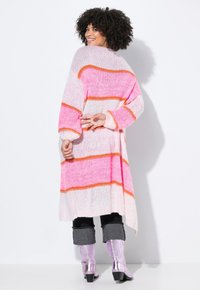 Strickjacke mit rosa und weißen horizontalen Streifen, mit einem orangen Akzent. Lange Ärmel, offenes Frontdesign und lockere Passform. Getragen über schwarzen Hosen.
