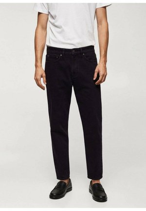 Homme portant un jean noir slim, des mocassins noirs et un t-shirt blanc uni à manches courtes, sur un fond neutre.