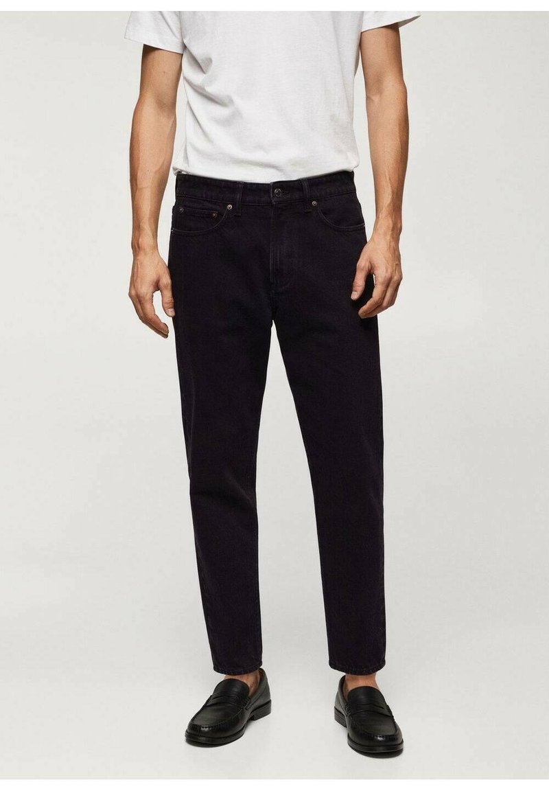 Homme portant un jean noir slim, des mocassins noirs et un t-shirt blanc uni à manches courtes, sur un fond neutre.