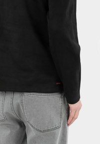 Maglietta nera a maniche lunghe con una texture liscia, caratterizzata da un piccolo logo rosso all'orlo, abbinata a jeans in denim grigio chiaro con tasche posteriori.