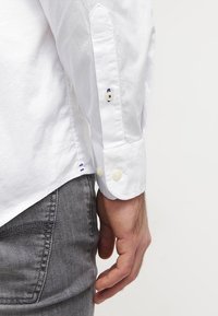 Witte overhemd met lange mouwen, voorzien van lichtblauwe en marineblauwe borduursels op de manchetten en witte knopen. Grijze jeans zichtbaar eronder.