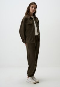 Olijfgroene, oversized jas met borstzakken en gouden knopen, gecombineerd met bijpassende joggers. Model draagt witte sneakers.