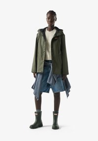 Modelo vistiendo una chaqueta impermeable color verde oliva, suéter crema, falda vaquera con camisa atada y botas de lluvia verdes a juego sobre fondo blanco.