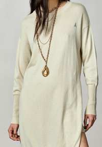 Vestido de punto color beige claro con mangas largas y aberturas laterales. Presenta un pequeño logo bordado en el pecho y una textura suave.
