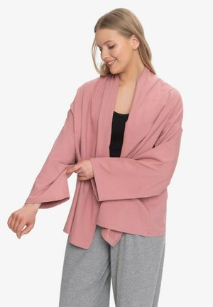 Uśmiechnięta kobieta poprawia rękaw luźnego, zakurzonego różowego kardiganu na czarnej bluzce, nosząc jasnoszare spodnie na białym tle.