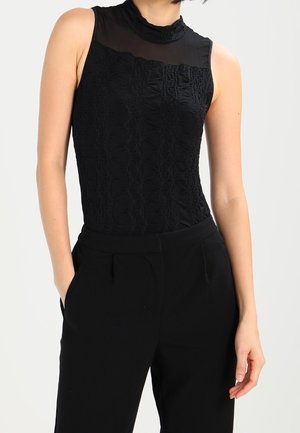 Kvinde iført sort ærmeløs bluse i blonder med gennemsigtig høj hals og sorte skræddersyede bukser, hænderne i lommerne, foran ensfarvet baggrund.