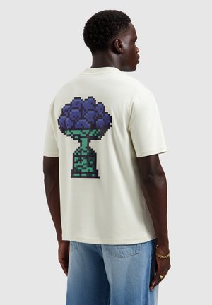 Man draagt een wit T-shirt met een gepixelde donkerblauwe bloemenboeket in een groene vaas op de rug, gecombineerd met lichtblauwe jeans.