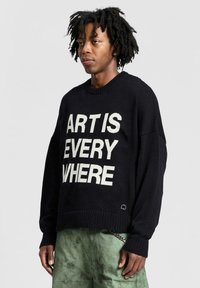 Maglione nero a maglia con testo bianco in bold che recita "L'ARTE È OVUNQUE", con un collo rotondo classico e polsini a coste.