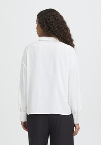 Hvid langærmet skjorte med et diskret prikmønster, blødt stof, afslappet pasform og knapper ved ærmerne, vist bagfra.