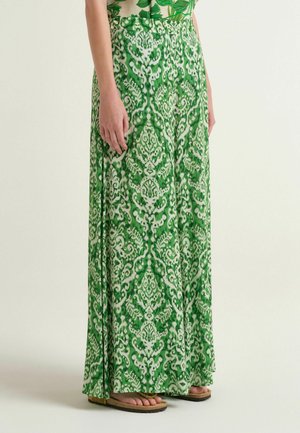 Gonna lunga con motivo verde e bianco, indossata con infradito, che mostra la parte inferiore del corpo di una persona dalla vita ai piedi su uno sfondo uniforme.