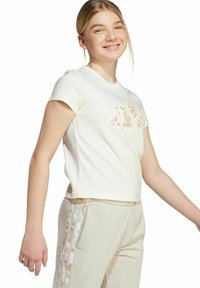 Jonge vrouw die glimlacht, gekleed in een witte cropped t-shirt met een panterprintlogo en lichtbeige broek met bijpassende panterprint zijkanten.