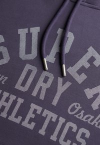 Paarse sweatshirt met grote grijze tekst "SUPER DRY ATHLETICS" en "Osaka." Voorzien van bijpassende trekkoorden met zilverkleurige metalen uiteinden.