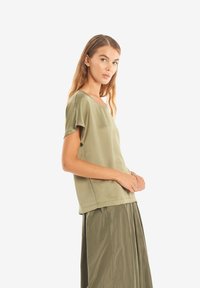 Top verde oliva a maniche corte realizzato in tessuto liscio; presenta una vestibilità rilassata e un collo a giro, abbinato a una gonna verde plissettata.