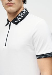 Άνδρας που φοράει λευκό πόλο με κοντά μανίκια, μαύρο φερμουάρ, μαύρο γιακά και μανσέτες στα μανίκια με την επωνυμία «Versace».