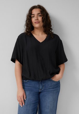 Vrouw met krullend haar die een losse zwarte V-hals blouse en blauwe jeans draagt, staand met één hand in haar zak tegen een effen muur.