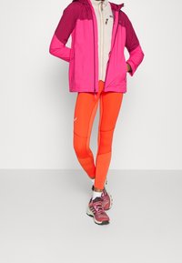 Heldere roze en paarse waterdichte jas met een capuchon, beige fleece eronder, gecombineerd met strakke oranje legging en roze sportschoenen.
