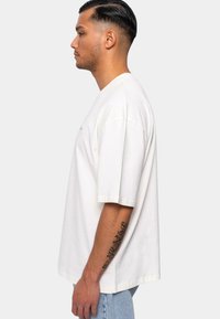 Overdimensioneret hvid bomulds T-shirt med korte ærmer og rund halsudskæring, med diskret broderi på brystet. Båret sammen med lyse denim jeans.