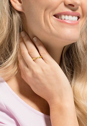 Goldring mit einer glatten Oberfläche, verziert mit einem kleinen dekorativen Punkt, getragen an einer Hand mit langen Fingern, neben einem hellrosa Oberteil.