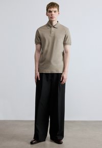 Polo cáqui com gola, mangas curtas e pequeno logótipo. Combinado com calças pretas largas e sapatos polidos. Tecido texturizado.