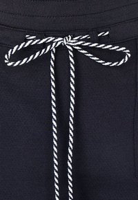 Tmavě modré tepláky s texturovaným povrchem, s bílým a tmavě modrým pleteným šňůrkem zavázaným v pase, viditelné detaily šití.