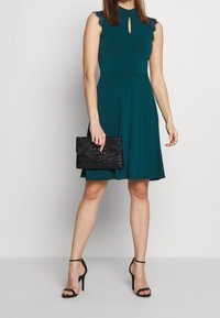 Robe bleu sarcelle avec encolure goutte d'eau et bordure en dentelle sur les épaules. Associée à une pochette noire texturée et des talons noirs à lanières.