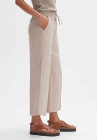 Pantalons larges en lin beige avec poches latérales et taille à cordon, arborant une coupe décontractée et des fentes à la cheville, associés à des sandales brunes.