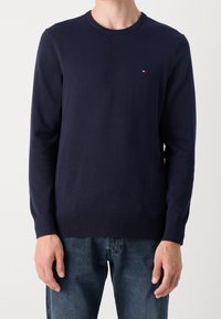 Pull en maille marine à manches longues, col rond et poignets côtelés. Présente un petit logo sur la poitrine. Le tissu semble doux et lisse.