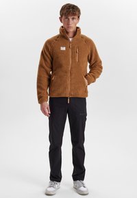 Brun fleecejakke med høj krave, lynlås foran og to sidelommer. Iført sorte bukser og hvide sneakers. Glat tekstur, afslappet design.