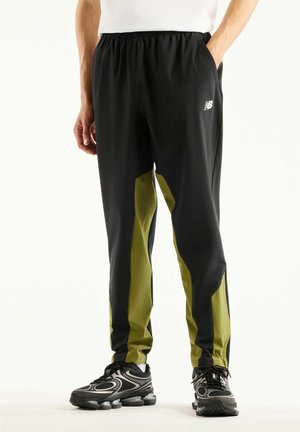 Man draagt zwarte en olijfgroene New Balance trainingsbroek en zwarte sportschoenen met zichtbare luchtkussenzolen.