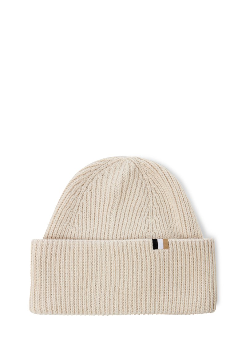 Knit beanie in light beige with a folded cuff. Features a textured rib pattern and a small brand tag in black, white, and tan at the side.