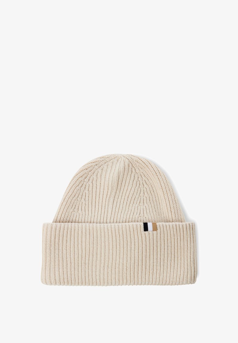 Knit beanie in light beige with a folded cuff. Features a textured rib pattern and a small brand tag in black, white, and tan at the side.