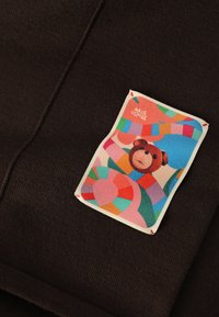 Zwarte textuurstof met een kleurrijk label met een berenafbeelding en abstracte vormen in pasteltinten, omrand met een roomkleurige rand.