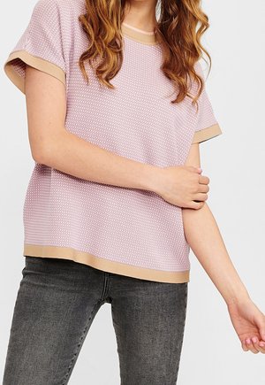 Kvinde iført en struktureret lyse lilla kortærmet top med beige kant og mørke grå jeans, stående med den ene arm krydset over kroppen.