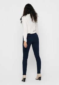 Blouse blanche à manches longues et pantalon ajusté bleu marine, avec une texture lisse et un profil élancé. Fini avec des chaussures à talons hauts de couleur claire.