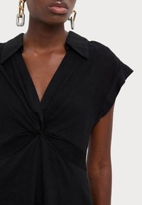 Svart skjortklänning med krage, korta ärmar och vriden knutdetalj i midjan, med ett mjukt, texturerat tyg.