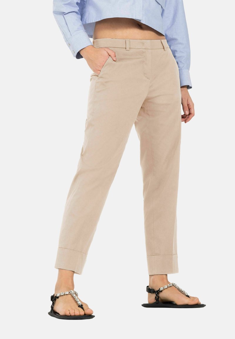 Beige katoenen broeken met een rechte snit, voorzien van zijzakken en opgerolde pijpen. Gecombineerd met zwarte sandalen met decoratieve banden.