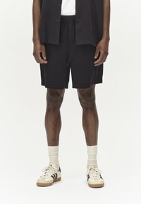 Schwarze Shorts mit einem elastischen Bund, kombiniert mit weißen Socken und beigen Sneakers mit schwarzen Streifen. Glatter Stoff, lockere Passform.