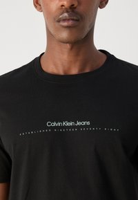 Juodas medvilnės marškinėliai su apvalia iškirpte ir spaudiniu, kuriame parašyta "Calvin Klein Jeans įsteigta 1978 metais."