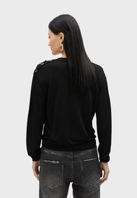 Svart tröja med långa ärmar, med guld detaljer vid axeln. Kombinerad med grå slitna jeans. Slät textur överlag.